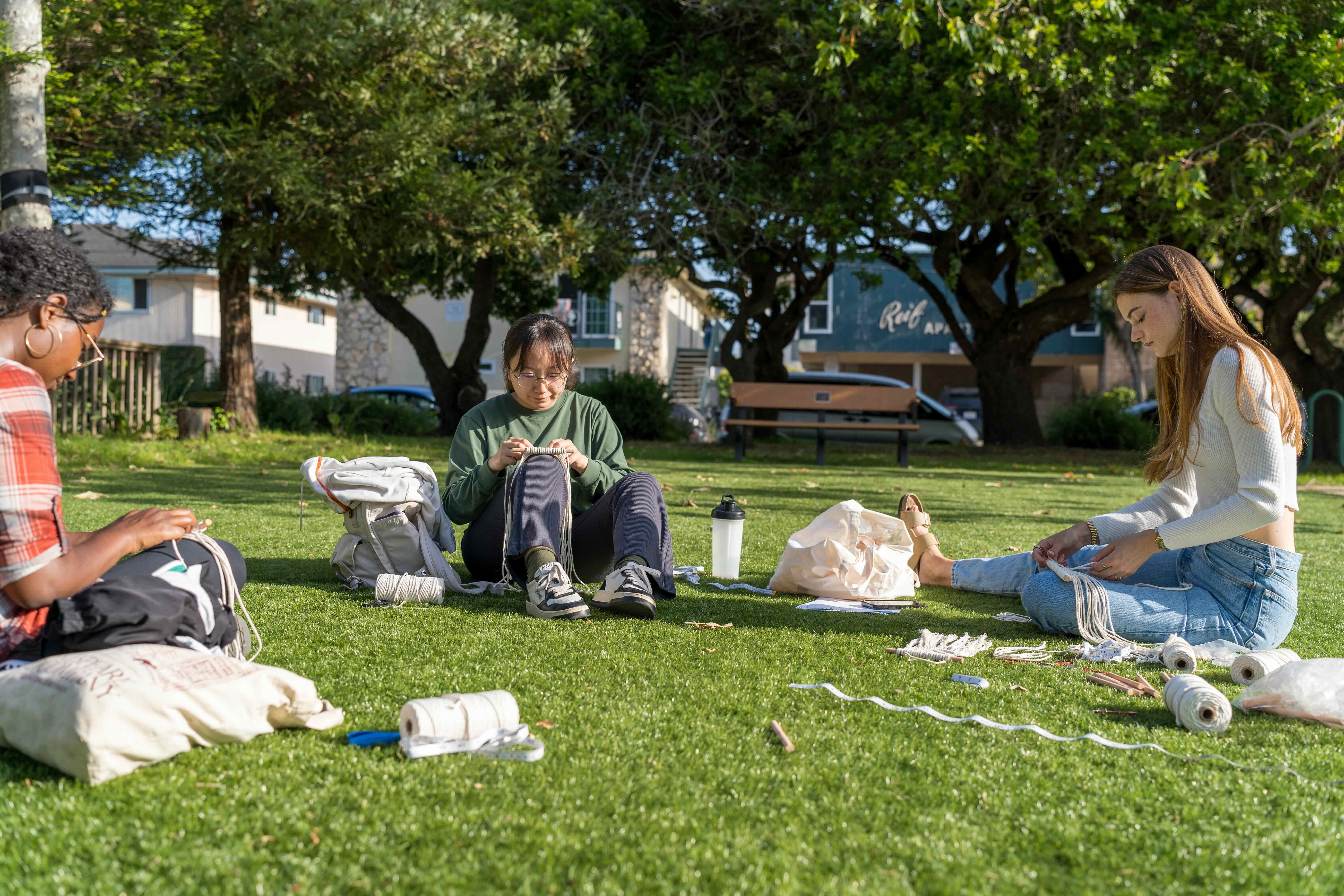 three girls sitting on the grass doing macrame 