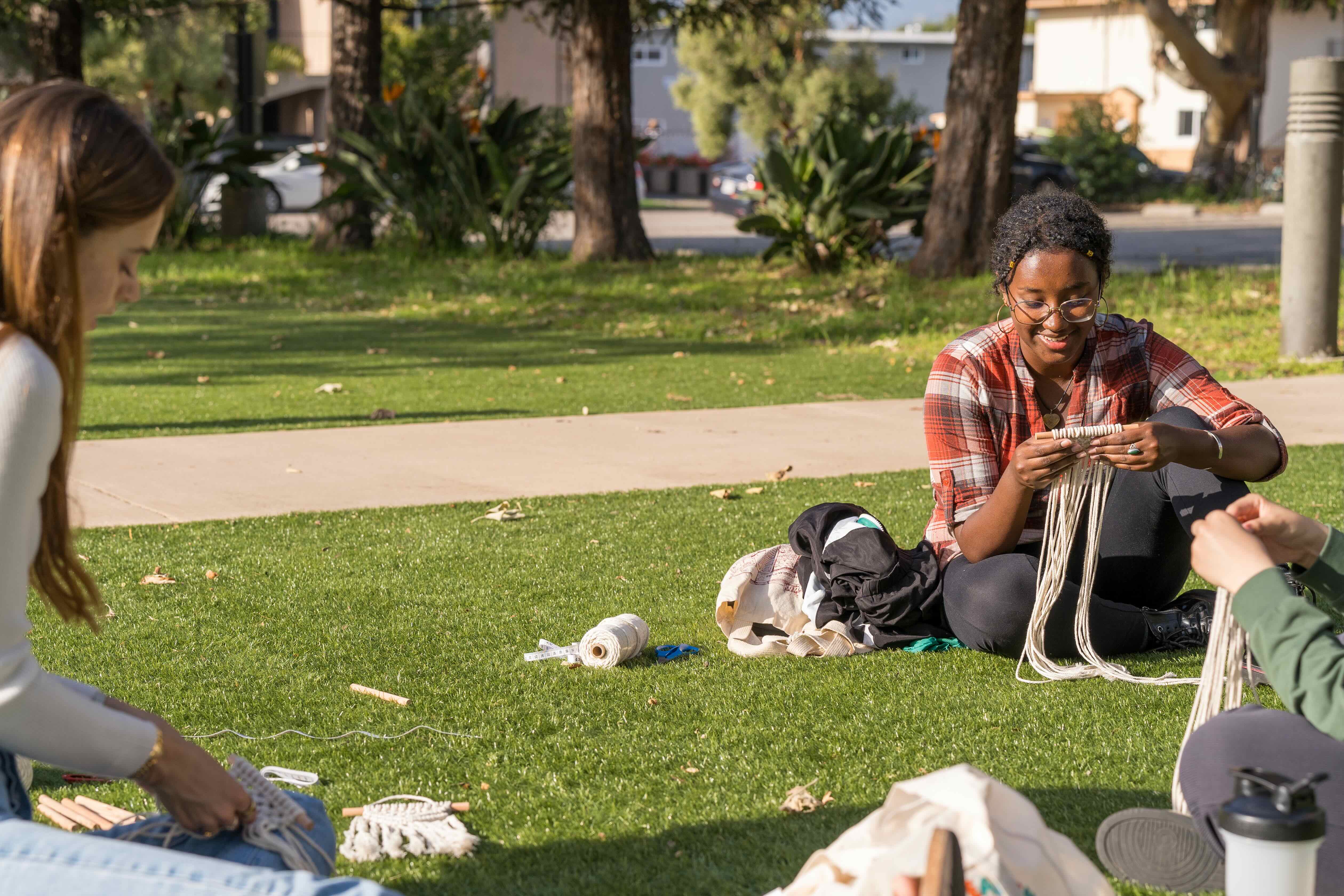 two girls sitting on the grass doing macrame 